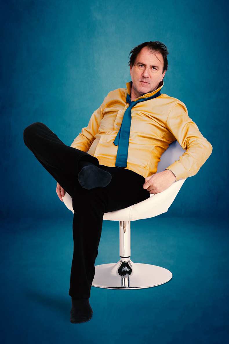 Stefan Waghubinger sitzt auf einem weißem Stuhl vor einem blauen Hintergrund und schaut in die Kamera.
