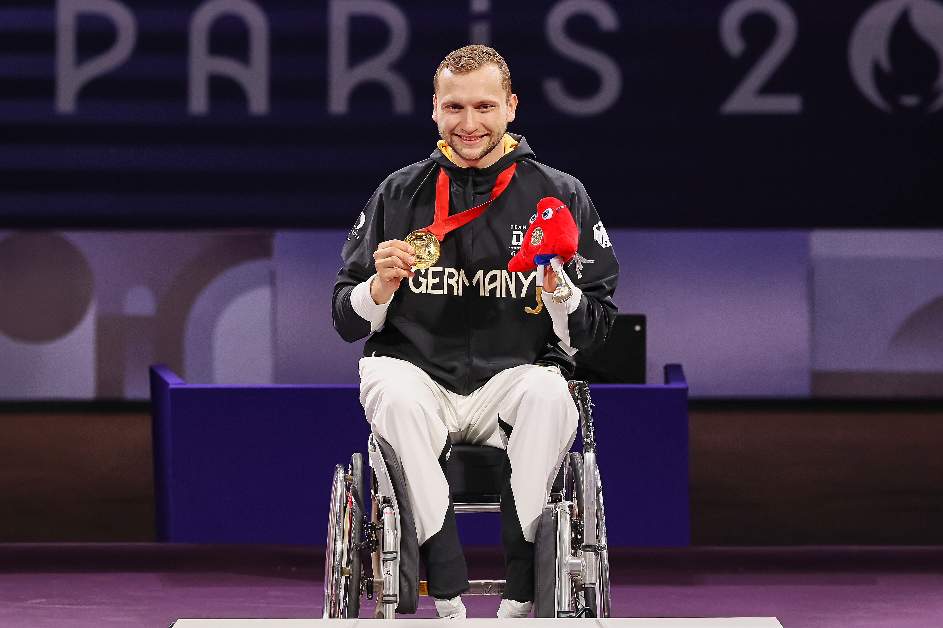 Rollstuhlsportler im Team-Germany-Outfit mit Goldmedaille und Maskottchen bei Siegerehrung.