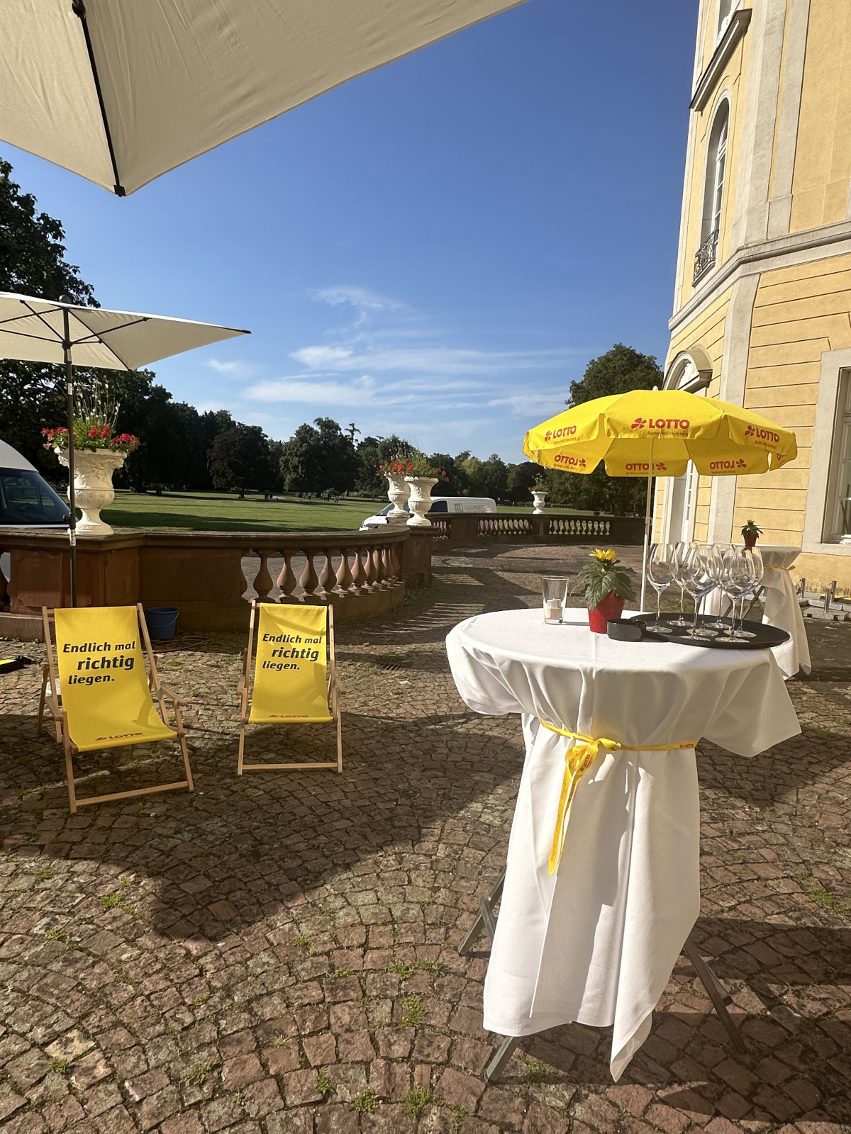 Lotto BW Liegestühle und Sonnenschirm mit Stehtisch auf Hof mit blauem Himmel
