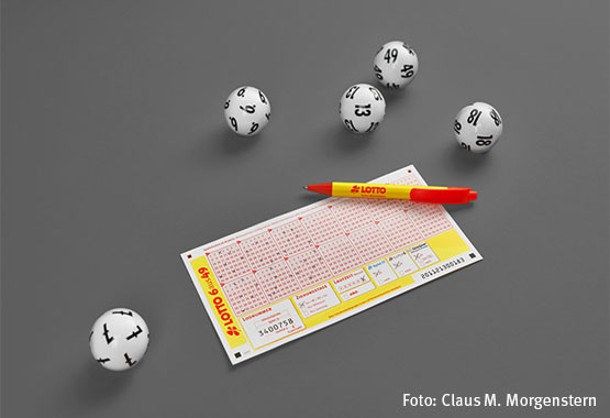 Lottoschein mit roten Zahlenfeldern, umgeben von weißen Kugeln und einem Lotto-Kugelschreiber auf grauem Hintergrund.