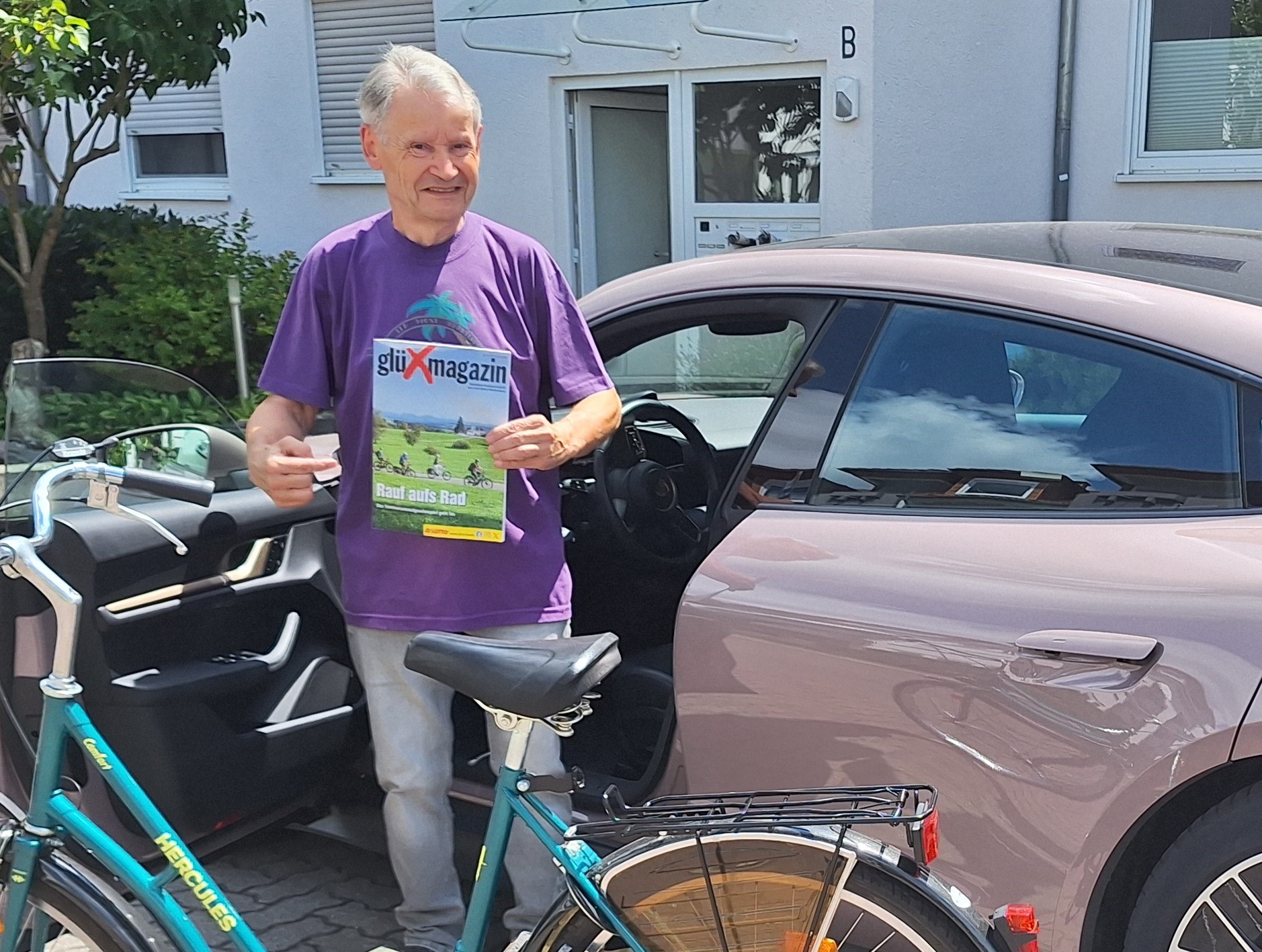 Mann mit Fahrrad und gl&uuml;Xmagazin-Ausgabe vor einem geparkten Auto.