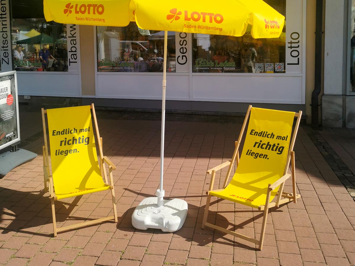 Im Freien stehen zwei Lotto-Liegestühle unter einem Lotto-Sonnenschirm