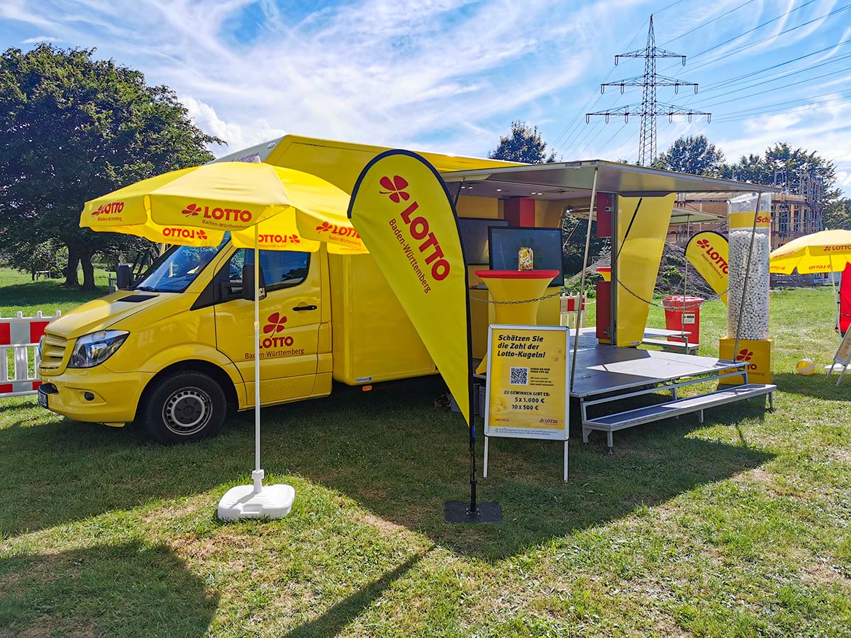 Das aufgeklappte Lotto-Mobil auf einer Wiese mit Sonnenschirm und Beachflags davor