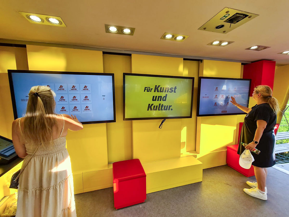 Drei Bildschirme an der Wand und zwei Frauen im Inneren des Lotto-Mobils