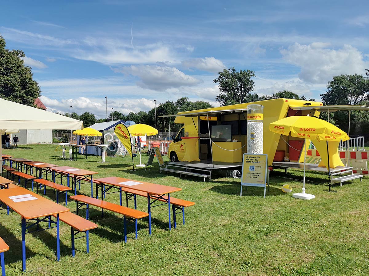 Das Lotto-Mobil auf einer Wiese bei Sonnenschein, davor stehen Tische und Bänke