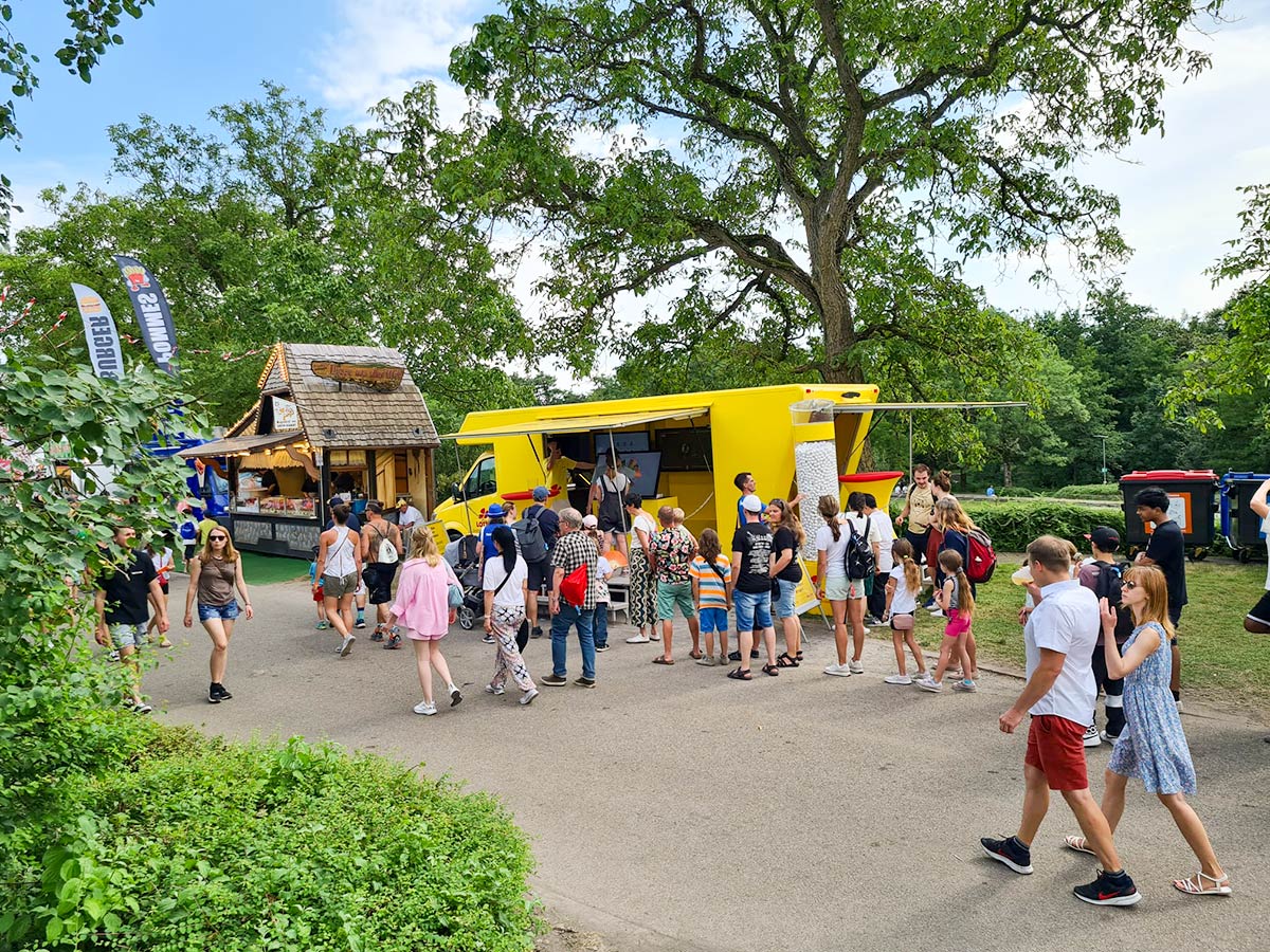 Das Lotto-Mobil am Wegrand in einem Park unter Bäumen mit zahlreichen Menschen, die daran vorbeilaufen