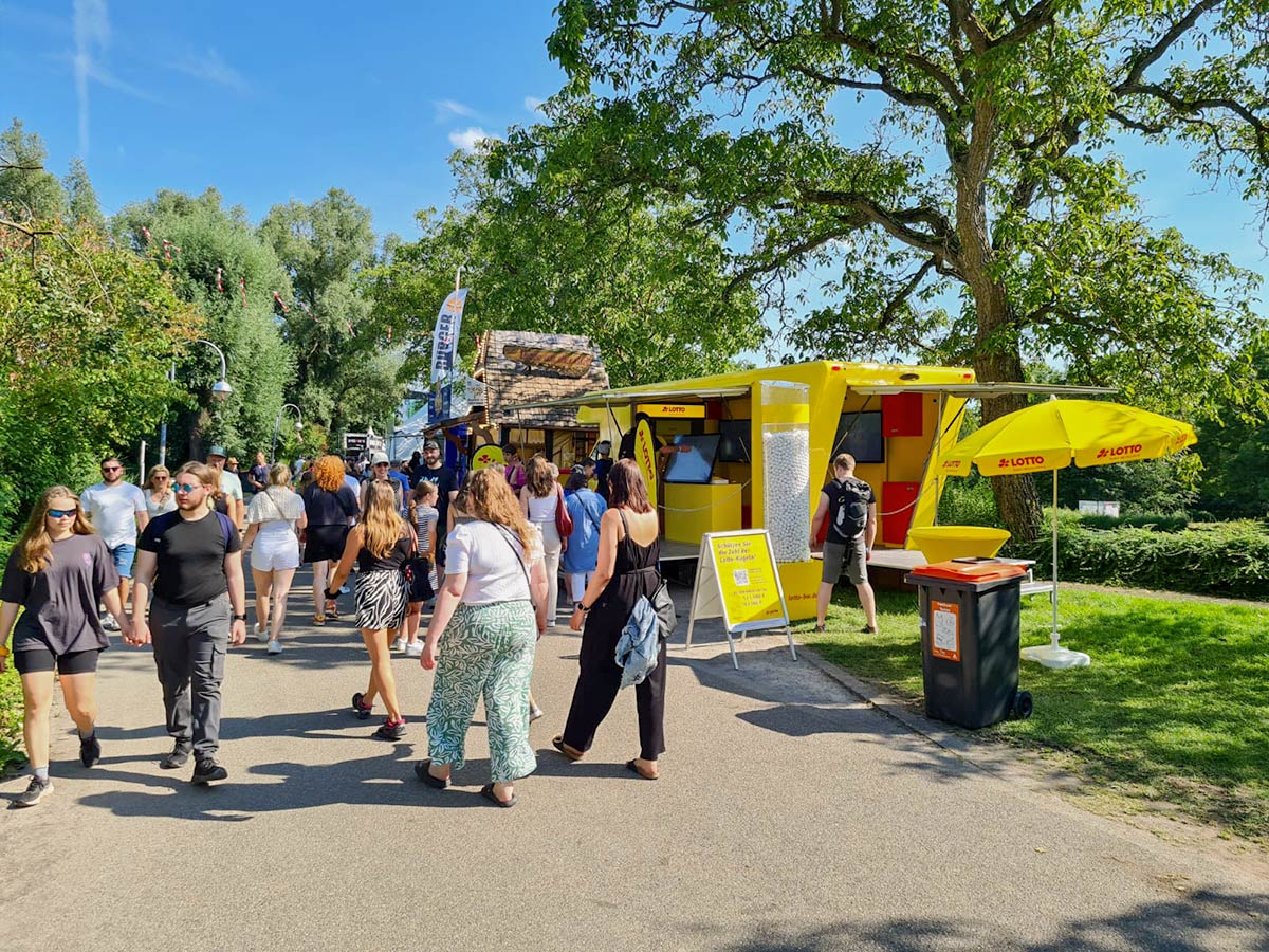 Das Lotto-Mobil am Wegrand in einem Park unter Bäumen mit zahlreichen Menschen, die daran vorbeilaufen