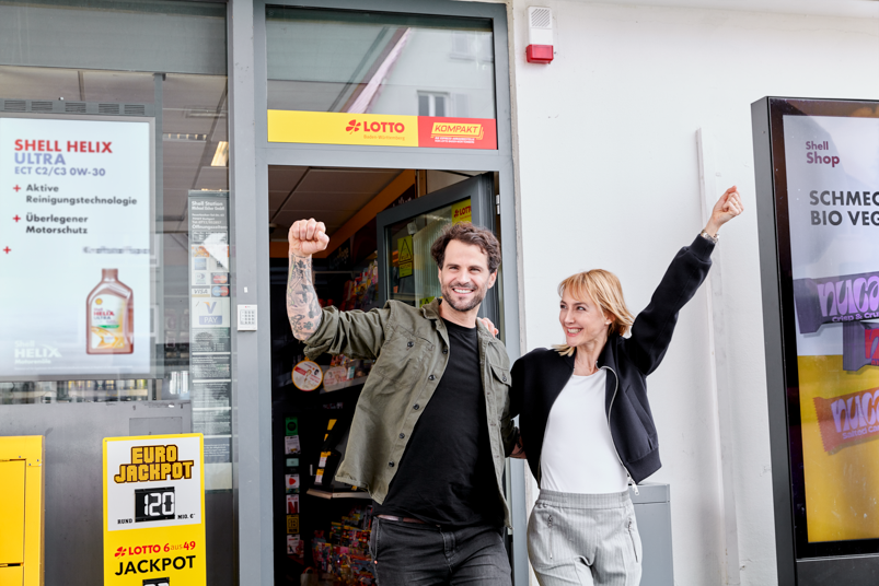 Paar vor einem Lotto-Shop, beide jubeln.