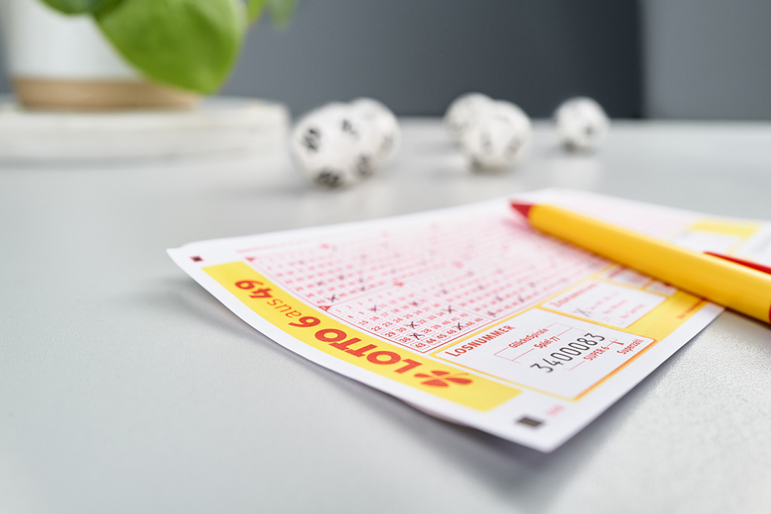 Spielschein und gelber Lotto-Stift liegen auf einem Schreibtisch. Im Hintergrund Lottokugeln.