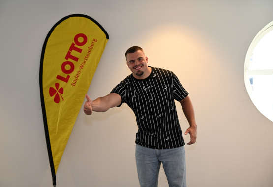Max Dehning lächelt neben einem Lotto BW Aufsteller in die Kamera