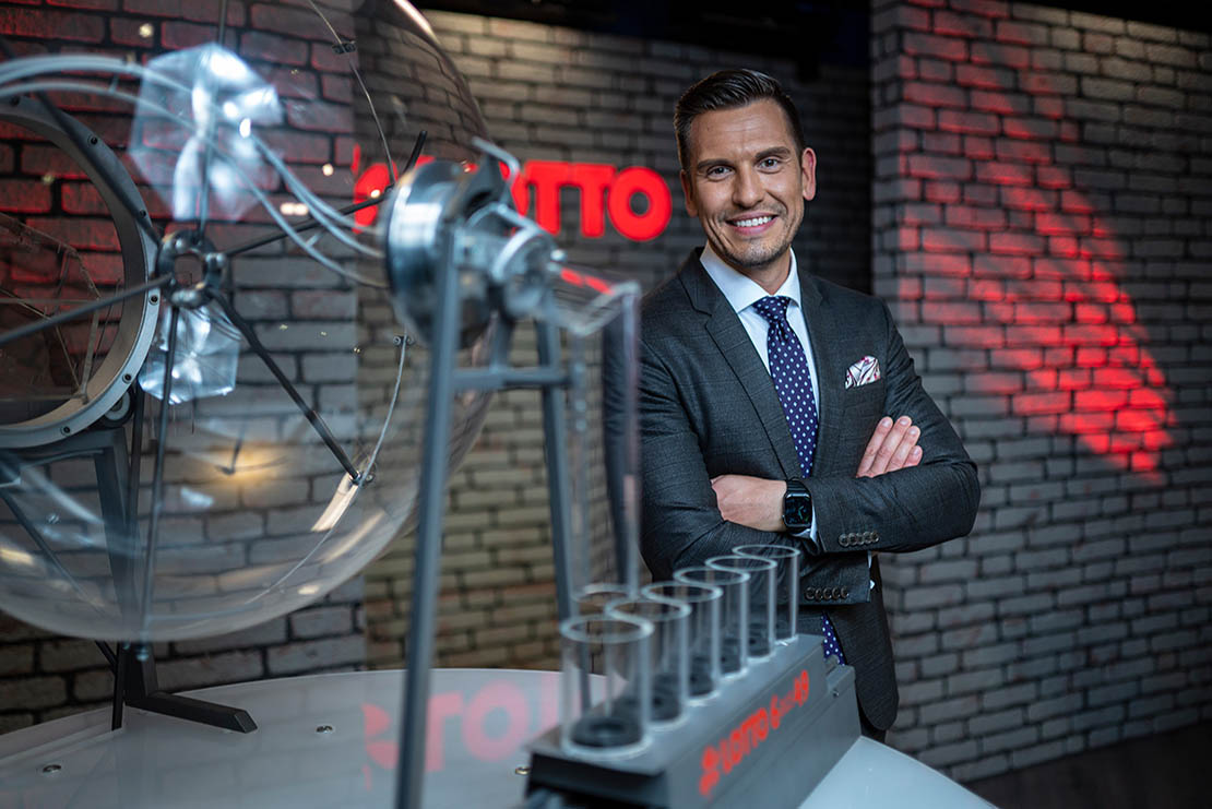 Moderator Chris Fleischhauer steht im Lotto-Studio neben einem Ziehungsgerät und lächelt in die Kamera.